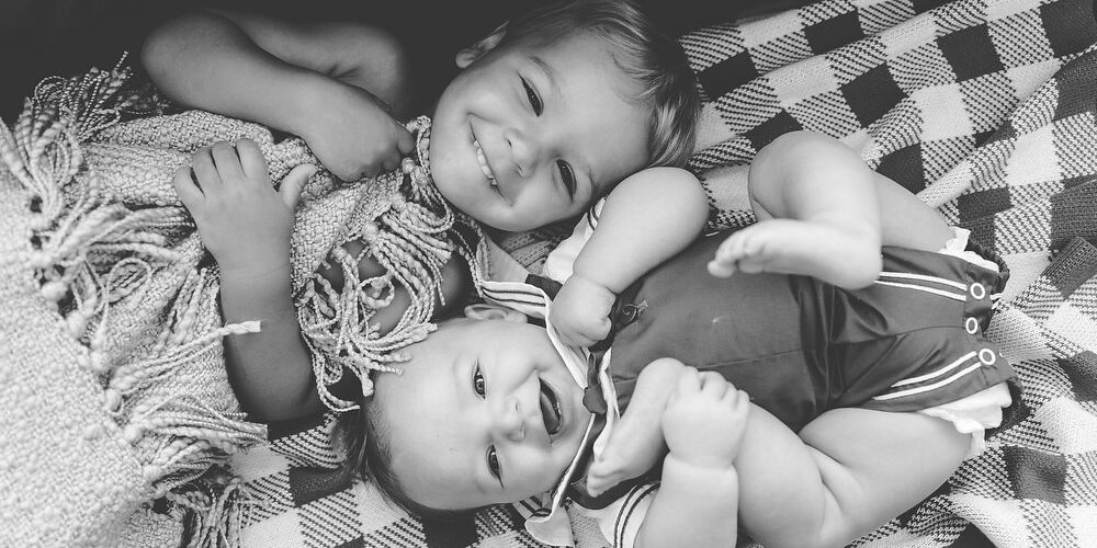 Two babies on a blanket smiling into the camera.
