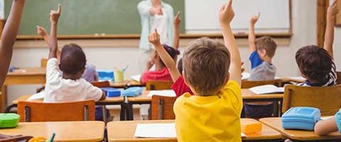 classroom-banner Classroom with children raising their hand with the view from back of room.