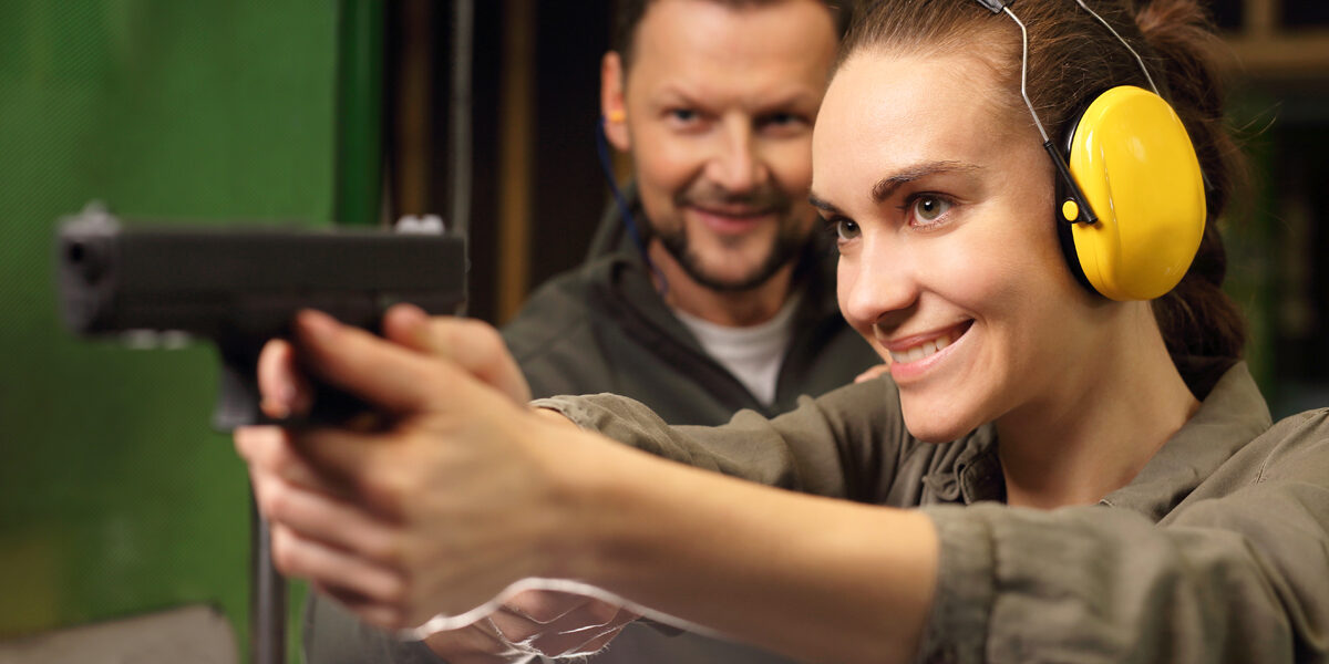 Woman shooting a gun at a inside a shooting range with a male instructor behind her.