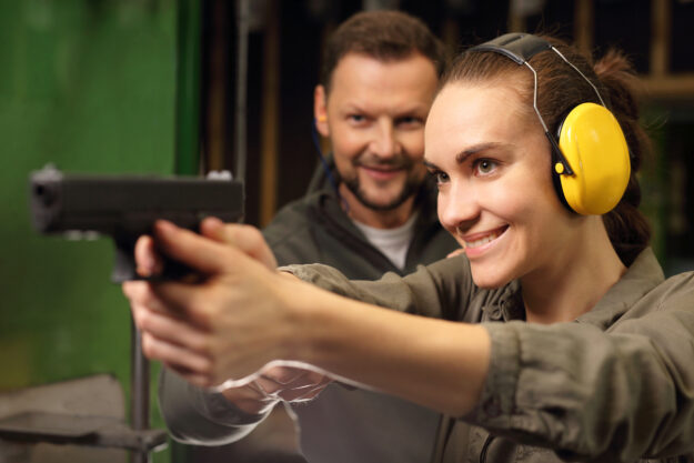 firearms-training-course Woman shooting a gun at a inside a shooting range with a male instructor behind her.