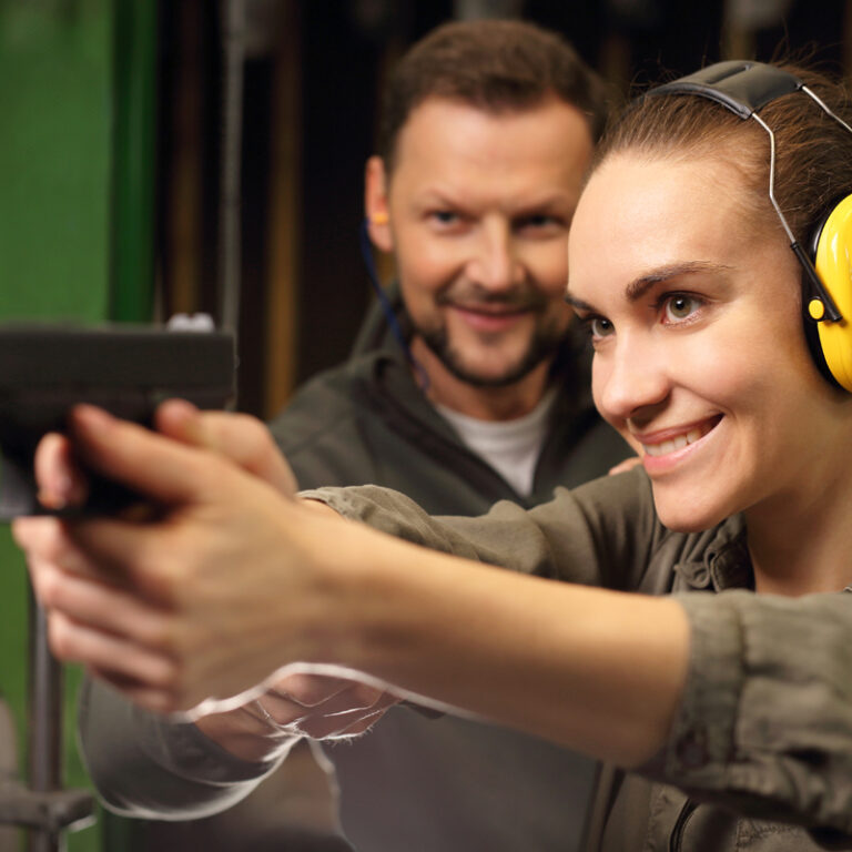 Woman shooting a gun at a inside a shooting range with a male instructor behind her.